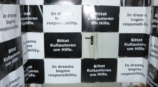 Die Fotografie zeigt eine große Metalltür, die mit schwarzen und weißen rechteckigen Papieren in einer Art Schachbrettmuster beklebt ist. Auf allen schwarzen Papieren ist in weißer Schrift der Text zu lesen: "Bittet Kultautoren um Hilfe". Auf allen weißen Papieren ist in schwarzer Schrift der Text zu lesen: "In dreams begins responsibility".