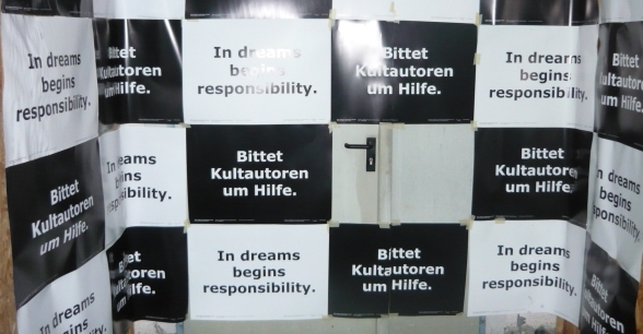 Die Fotografie zeigt eine große Metalltür, die mit schwarzen und weißen rechteckigen Papieren in einer Art Schachbrettmuster beklebt ist. Auf allen schwarzen Papieren ist in weißer Schrift der Text zu lesen: "Bittet Kultautoren um Hilfe". Auf allen weißen Papieren ist in schwarzer Schrift der Text zu lesen: "In dreams begins responsibility".