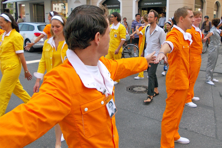 Das Bild zeigt Performer*innen, die in gelbe, orangene und silberne Overalls gekleidet sind, auf der Dienerstraße in der Münchener Innenstadt. Einige Performer*innen halten sich an den Händen und bilden zwei Menschenketten, in deren Mitte ein Gang gebildet wird. Durch dieses Gang laufen weitere Performer*innen sowie Passant*innen.
