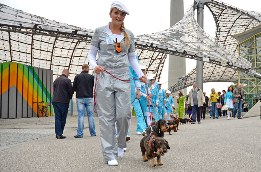 Das Bild zeigt eine im Olympiapark aufgenommene Fotografie. Auf dem Coubertinplatz vor der Olympiaschwimmhalle laufen eine Gruppe von Performer*innen in einer Reihe, sie sind alle in einfarbige Overalls in Blau, Silber und Grün sowie weiße Schirmmützen gekleidet. Alle Performer*innen führen jeweils einen Dackel an der Leine und bilden damit eine Dackelparade. Die Parade wird begleitet von einer Reihe von Passant*innen und Schaulustigen.