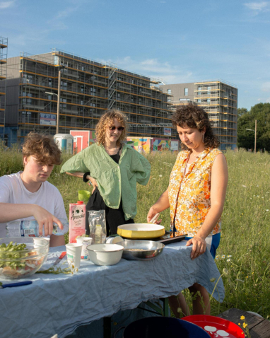 Drei Personen stehen und sitzen an einem provisorischen Tisch im Freien und bereiten Essen zu. Im Hintergrund sind hohe, im Bau befindliche Gebäude zu sehen.