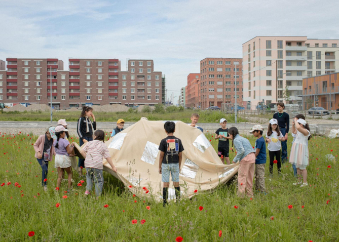 einige Kinder und zwei Erwachsene stehen im Kreis in einer Wiese und halten ein beiges Tuch, im Hintergrund Häuserblocks