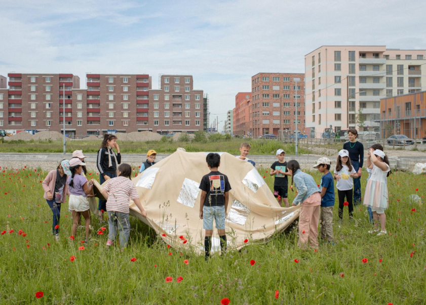 einige Kinder und zwei Erwachsene stehen im Kreis in einer Wiese und halten ein beiges Tuch, im Hintergrund Häuserblocks