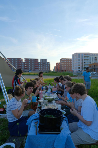 Mehrere Menschen sitzen an einem gedeckten Tisch im Freien und essen, auf dem Tisch steht ein großer Topf, Schüsseln und Flaschen. Im Hintergrund stehen Neubauten und Baustellen.