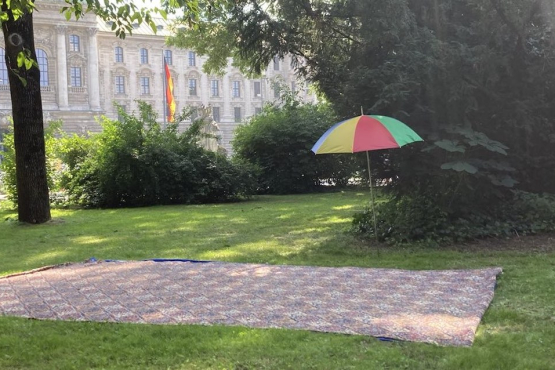 Fotografie einer Grünflache im Alten Botanischen Garten. Auf dem Rasen ist eine große teppichartige Decke ausgelegt, daneben steht eine Sackkarre sowie ein aufgespannter, bunter Sonnenschirm. Im Hintergrund zu sehen ist der Justizpalast.
