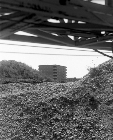 In Schwarz-Weiß zeigt das Bild einen mehrstöckigen Wohnblock in der Ferne, umgeben von Hügeln aus Schutt oder Geröll. Im Vordergrund sind unscharfe Metallstreben und Geröll zu sehen, die den Blick auf das Gebäude teils verdecken.