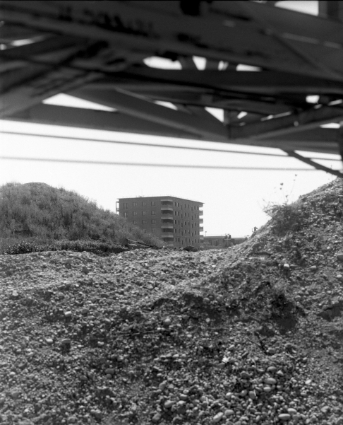 In Schwarz-Weiß zeigt das Bild einen mehrstöckigen Wohnblock in der Ferne, umgeben von Hügeln aus Schutt oder Geröll. Im Vordergrund sind unscharfe Metallstreben und Geröll zu sehen, die den Blick auf das Gebäude teils verdecken.
