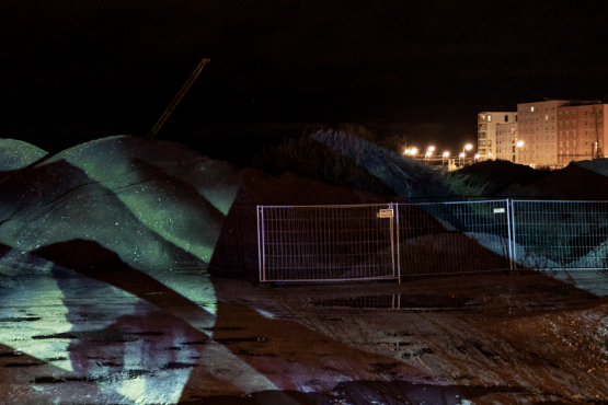 Das Bild zeigt eine Baustelle bei Nacht mit abgesperrten Erdhaufen, auf die eine abstrakte Lichtprojektion geworfen wird. Im Hintergrund sind beleuchtete Wohngebäude zu sehen.