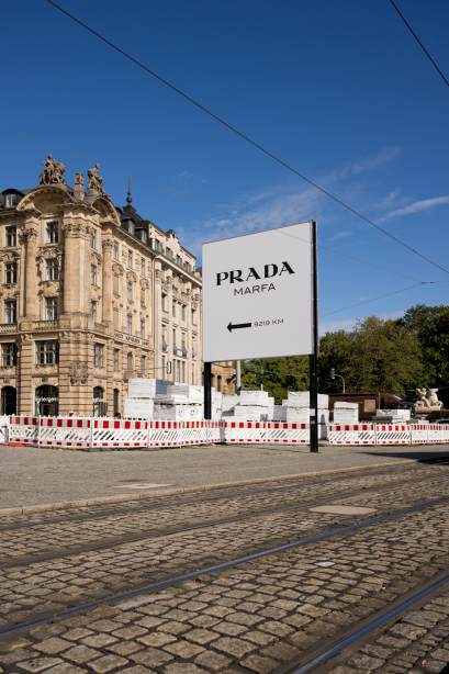 Das Billboard mit dem Motiv von Elmgreen und Dragset zeigt den Text "Prada Marfa 9219km" mit einem Pfeil, der von rechts nach links zeigt. Das Billboard steht vor einem historischen Gebäude in einer Stadt.