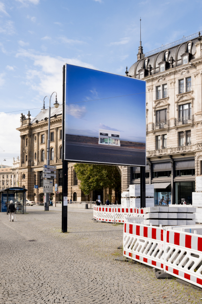Das Billboard mit dem Motiv von Elmgreen und Dragset zeigt eine einsame Prada-Boutique in einer Wüstenlandschaft. Das Billboard steht vor einem historischen Gebäude in einer Stadt.