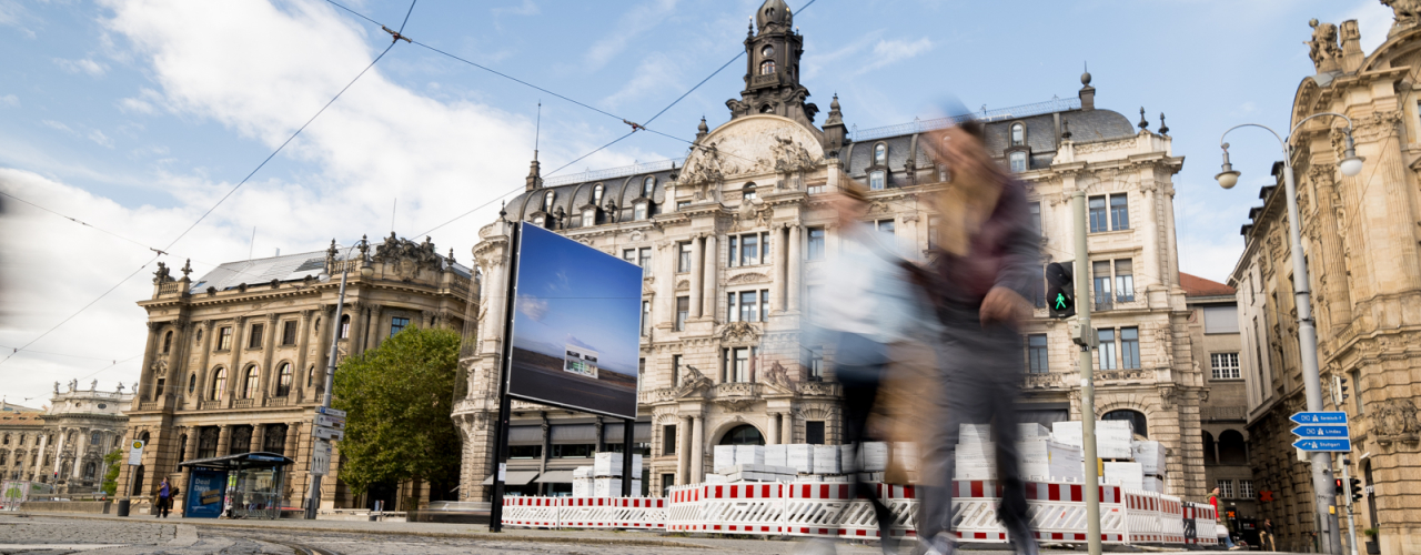 Das Billboard mit dem Motiv von Elmgreen und Dragset zeigt eine einsame Prada-Boutique in einer Wüstenlandschaft. Das Billboard steht vor einem historischen Gebäude in einer Stadt, während zwei Personen unscharf im Vordergrund vorbeigehen.