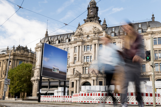 Das Billboard mit dem Motiv von Elmgreen und Dragset zeigt eine einsame Prada-Boutique in einer Wüstenlandschaft. Das Billboard steht vor einem historischen Gebäude in einer Stadt, während zwei Personen unscharf im Vordergrund vorbeigehen.