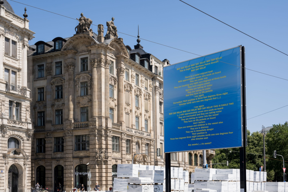 Das Billboard zeigt einen Notruftext in gelber Schrift vor blauem Hintergrund. Dahinter sind historische Gebäude mit verzierten Fassaden zu sehen. Im Vordergrund liegen gestapelte Kisten.