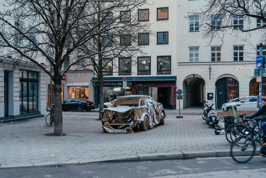 Ein SUV aus Lehm steht an einer Straßenkreuzung in einer städtischen Umgebung. Der SUV zeigt erste Zerfallserscheinungen und ist leicht mit Schnee bedeckt.