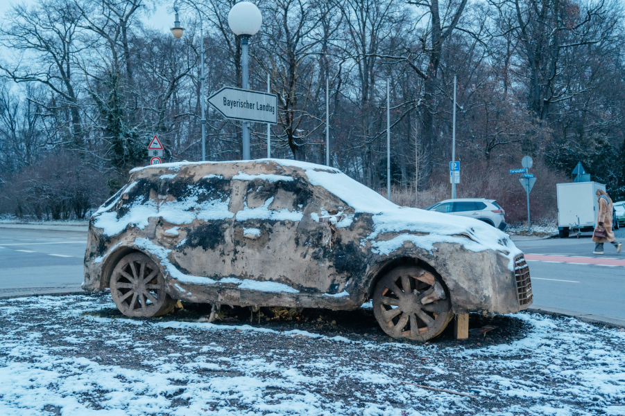 Ein SUV aus Lehm steht auf einer Verkehrsinsel in einer städtischen Umgebung, umgeben von Gebäuden und Straßenlaternen. Alles ist leicht mit Schnee bedeckt.