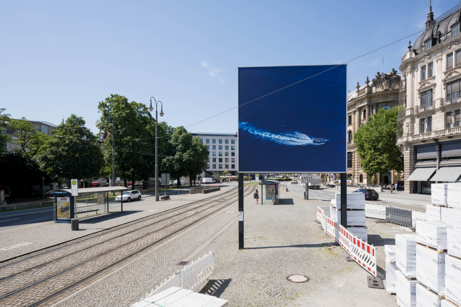 Das Billboard zeigt ein Luftbild eines Schnellboots auf offenem Meer. Es steht an einer Straßenbahn-Haltestelle in einer städtischen Umgebung mit historischen Gebäuden, Bäumen und gestapelten Kisten im Vordergrund.