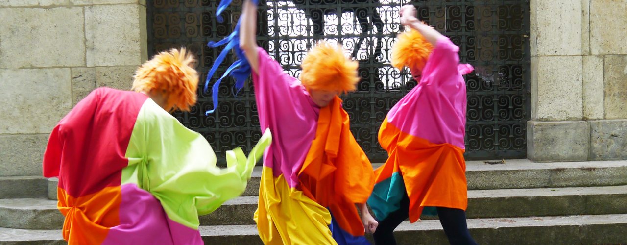 3 people with orange hair in colourful costumes move wildly in front of steps and a barred gate.