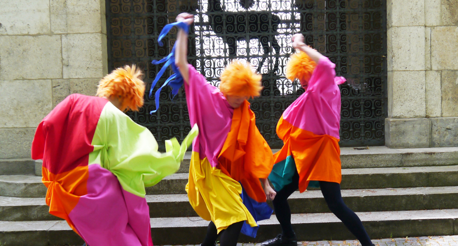 3 people with orange hair in colourful costumes move wildly in front of steps and a barred gate.