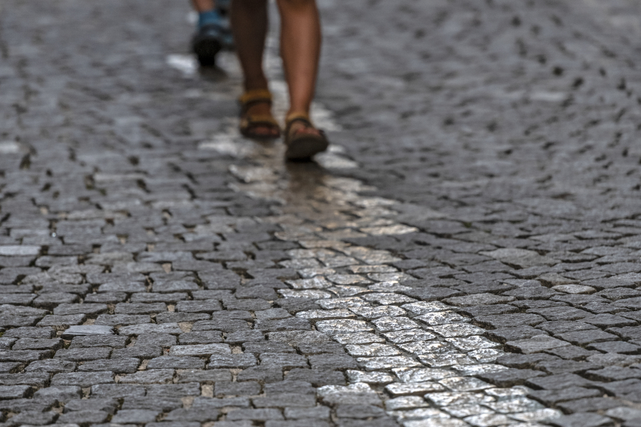 Detailaufnahme einer gepflasterten Fläche, ein Streifen mit einer Breite von ein bis vier Steinen hebt sich durch eine gold-glänzende Oberfläche ab, dieser Streifen wird von zwei Personen beschritten, von denen fast nur die Füße zu sehen sind.