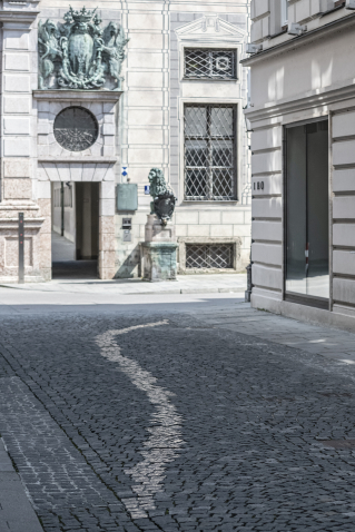 Blick auf eine gepflasterte Straße vor einer historischen Häuserwand. Ein geschlängelter Streifen glänzender Pflastersteine hebt sich deutlich ab.