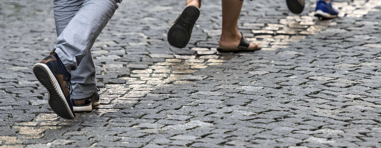 Detailaufnahme einer gepflasterten Fläche, ein Streifen mit einer Breite von ein bis vier Steinen hebt sich durch eine gold-glänzende Oberfläche ab, dieser Streifen wird von drei Personen beschritten, von denen fast nur die Füße zu sehen sind.