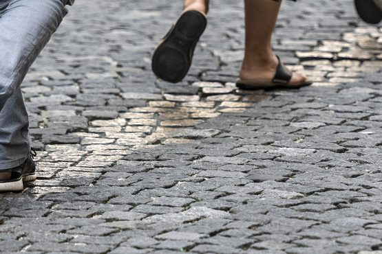 Detailaufnahme einer gepflasterten Fläche, ein Streifen mit einer Breite von ein bis vier Steinen hebt sich durch eine gold-glänzende Oberfläche ab, dieser Streifen wird von drei Personen beschritten, von denen fast nur die Füße zu sehen sind.