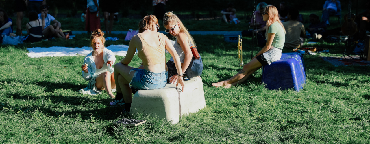 Two bench-like objects in beige and purple, standing in the grass, people dressed in summer clothes sitting on them or on the ground, blankets lying in the background.
