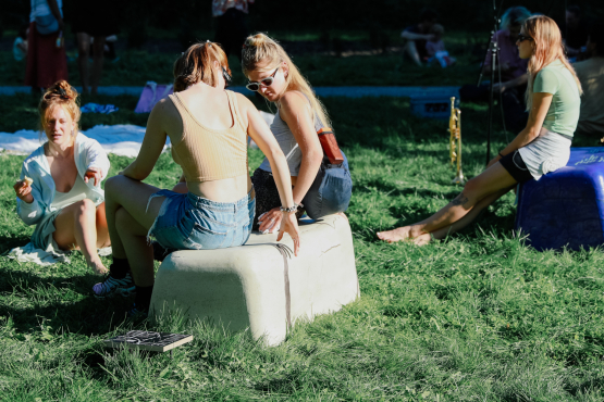 Two bench-like objects in beige and purple, standing in the grass, people dressed in summer clothes sitting on them or on the ground, blankets lying in the background.