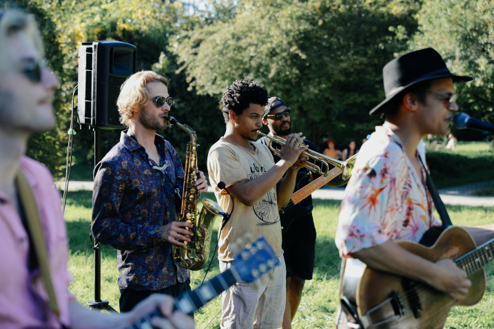 Several people play various instruments such as trumpet, saxophone and guitar in a meadow, with trees in the background.