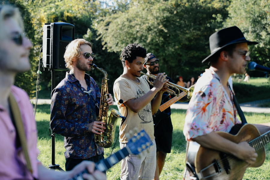 Several people play various instruments such as trumpet, saxophone and guitar in a meadow, with trees in the background.