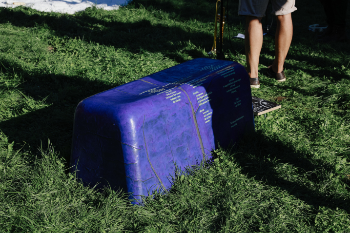 Purple bench-like object with yellowish lettering stands in the grass, next to it legs of people.