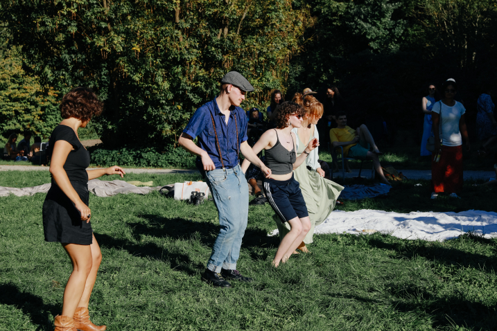 Several summery dressed people dance barefoot in the grass, blankets lie in the background.