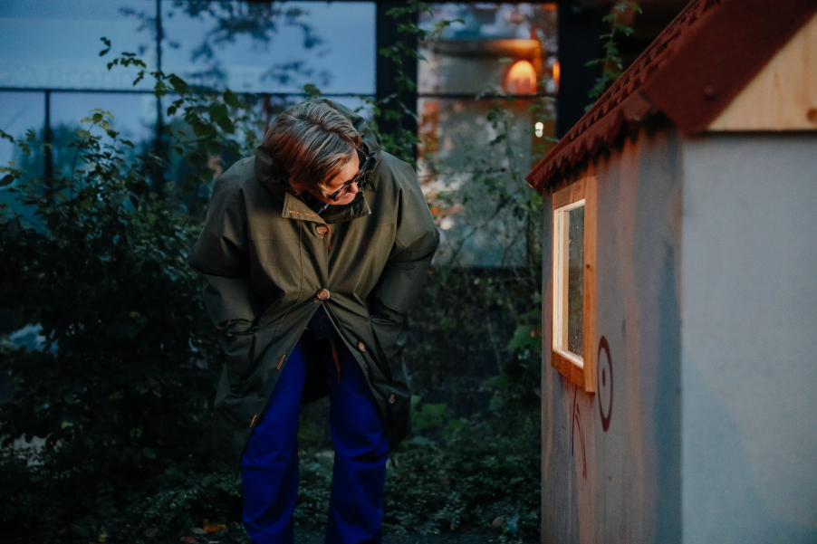 Person bends down in the twilight to a small house and looks in the window.