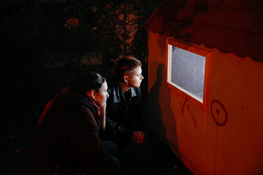 Two people look through a window in the dark, their faces illuminated by the light from inside.