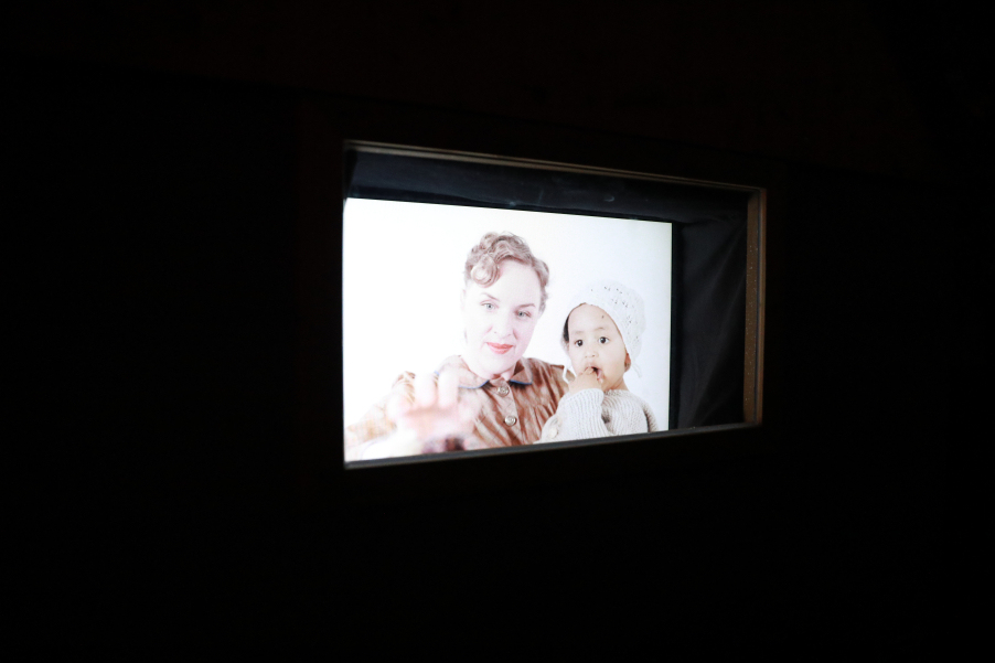 Oblique view of a screen in a dark room, showing a blonde woman holding a child with darker skin and brown eyes.