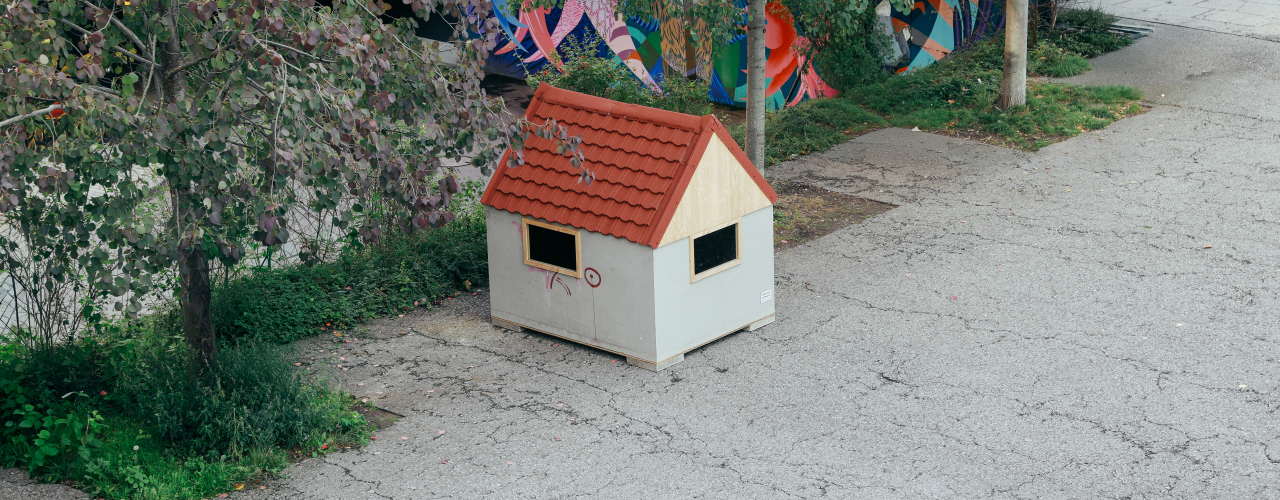 A very small house, seen from diagonally above, stands on an asphalt surface, with a strip of meadow behind it.