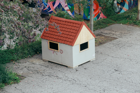 A very small house, seen from diagonally above, stands on an asphalt surface, with a strip of meadow behind it.