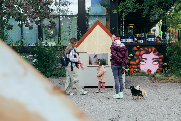 Frontal view of a small house, in front of it a woman with a baby, a woman with two small dogs and a child.