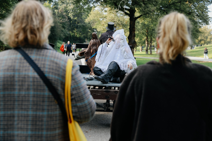 A carriage drives through a park, on the carriage stands a brownish Madonna sculpture and sits a person covered in white lace holding a microphone.