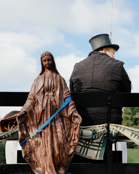 Frontal view of a large sculpture of the Madonna made of brown material in front of a slightly cloudy sky, with a person dressed in black and wearing a top hat sitting behind it.