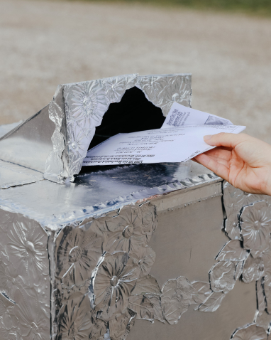 A hand inserts a letter into a letterbox covered with silver foil embossed with flowers.