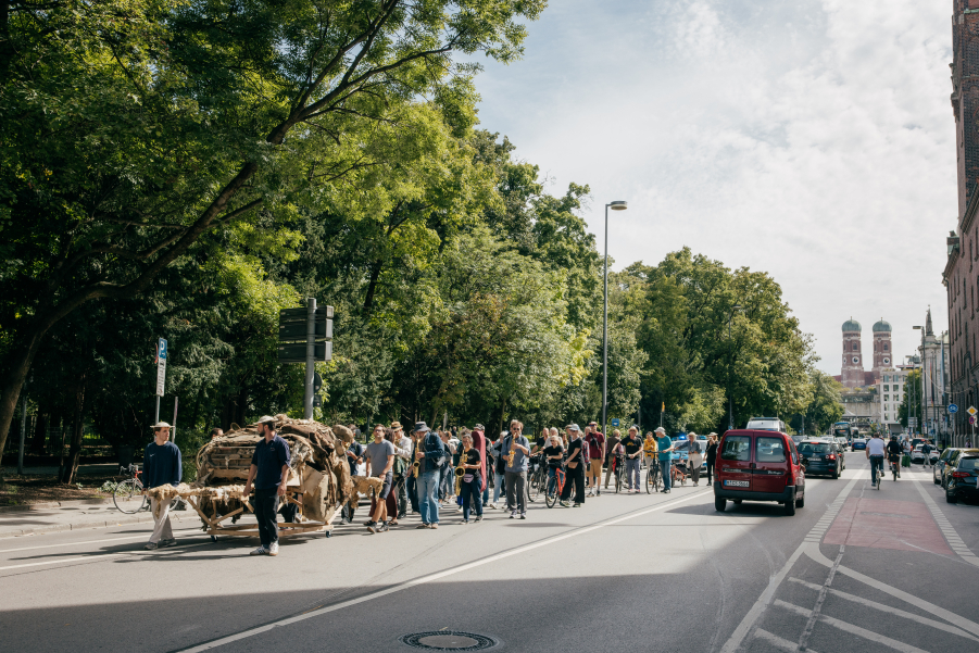 Mehrere Personen schieben ein kunstvolles Objekt, Überreste des zerfallenen SUVs aus Lehm, durch eine Straße, gefolgt von einigen Menschen. Im Hintergrund sind die Türme der Münchner Frauenkirche zu sehen.
