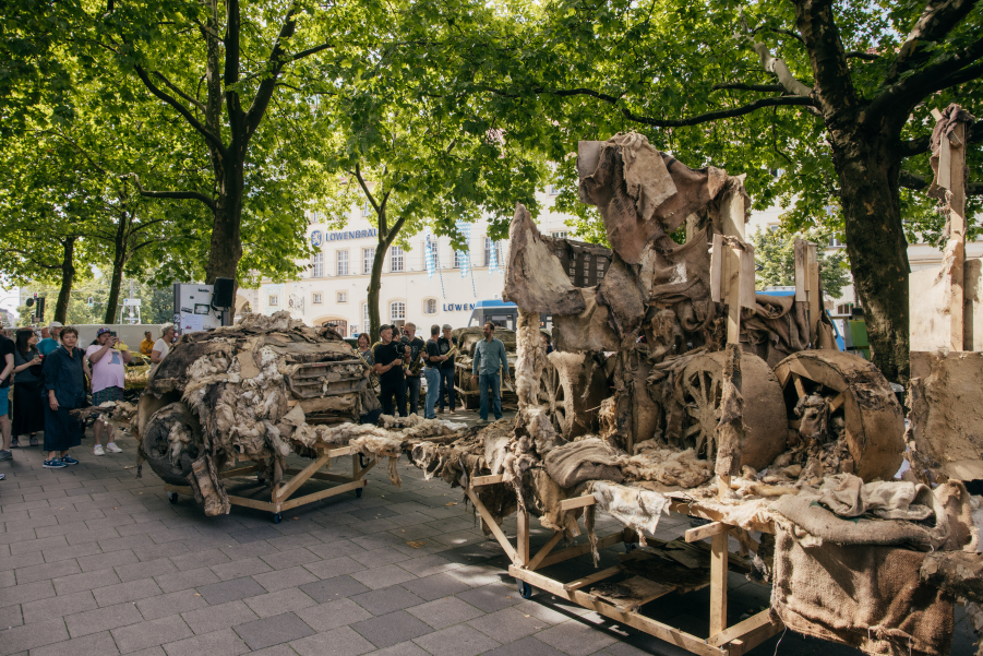 Überreste der zerfallenen SUVs aus Lehm auf Holz-Gerüsten stehen auf einem Platz unter Bäumen, einige Menschen stehen drumrum.