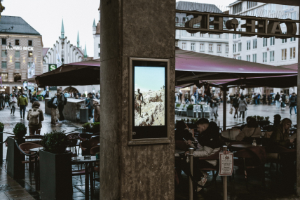 Vitrine an einem Pfeiler mit einem digitalen Bildschirm, auf diesem ist eine Person in einer Felslandschaft zu sehen. Im Hintergrund ein Straßencafé und belebte Stadtlandschaft.