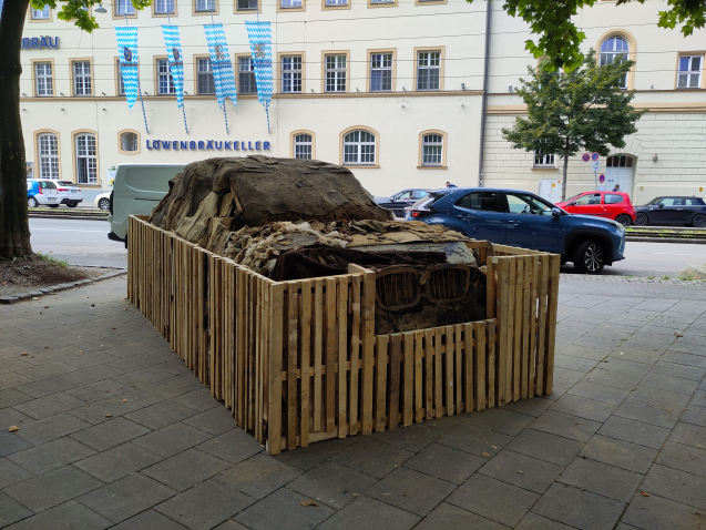Der zerfallene SUV aus Lehm ist mit einem Bretterzaun eingezäunt, im Hintergrund sieht man ein großes gebäude mit der Aufschrift 'Löwenbräukeller'