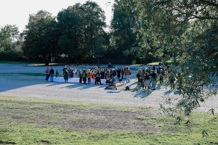Outdoors, many people are standing on a gravel area around a long table covered with white cloths. Trees in the background.
