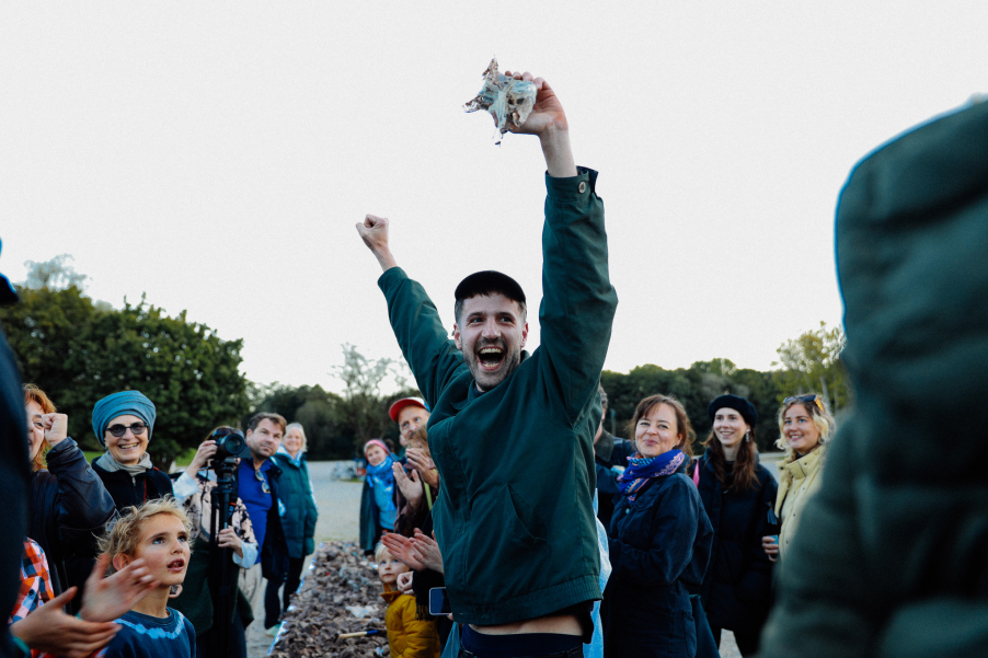 A person holds a broken fragment in the air jubilantly, surrounded by applauding and laughing people outdoors.