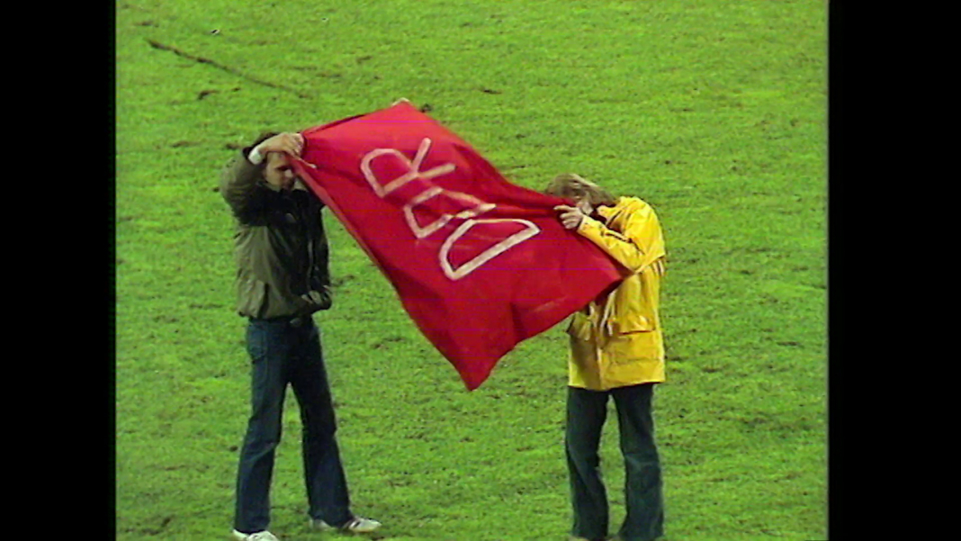 Zwei Personen auf einer Rasenfläche, sie halten ein rotes Tuch hoch auf dem DDR steht.