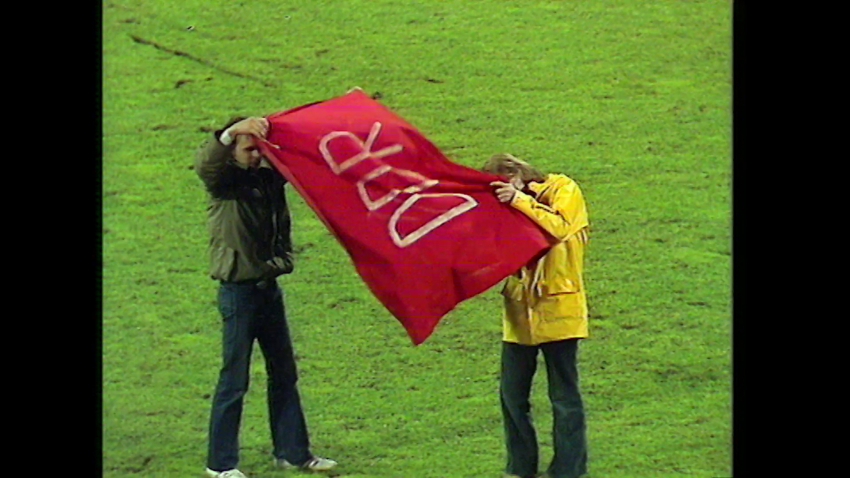 Zwei Personen auf einer Rasenfläche, sie halten ein rotes Tuch hoch auf dem DDR steht.