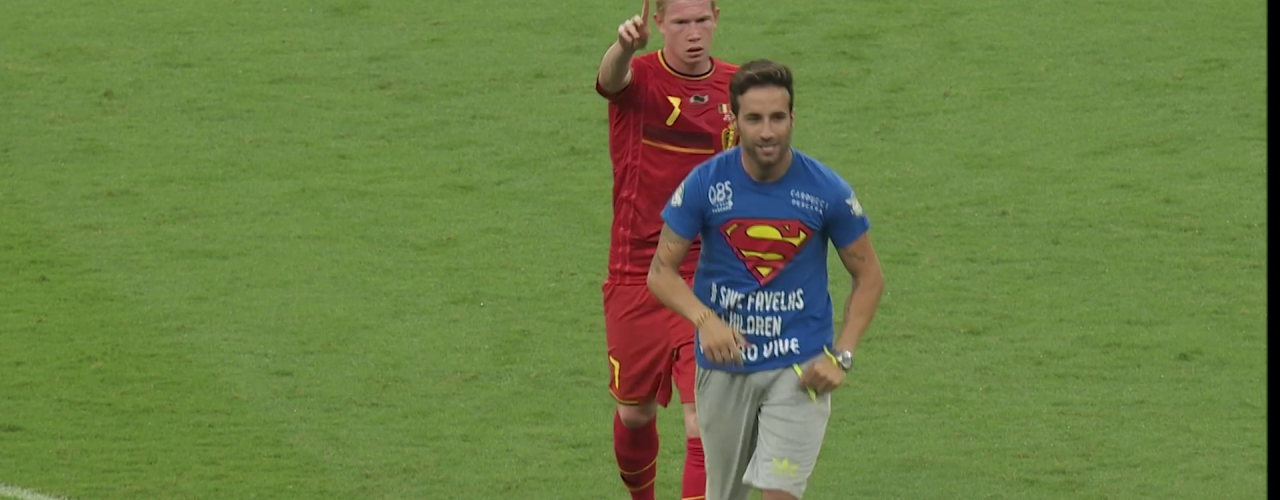 Fußballspieler im roten Trikot zeigt mit dem Finger nach vorn, vor ihm läuft ein Mann im blauen T-Shirt über den Rasen.